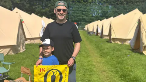 Ronan Cooper and his son, Alex, are seen at the campsite holding a yellow flag that has a number of signatures on it.