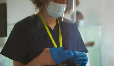 A woman with brown curly hair, wearing blue gloves, a yellow lanyard, a face visor and a blue medical scrubs (generic image).