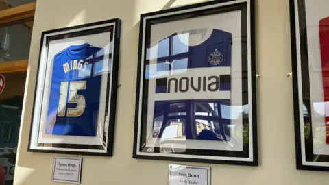 Various signed sports tops and t-shirts in frames on a white wall. Below them are names with information.