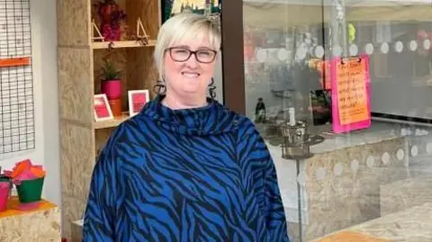 Becky Carrier in a blue animal print top standing in font of market stall called The Creative Place