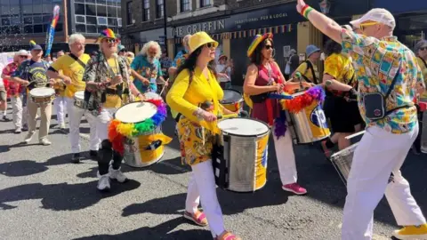 A Pride parade with men and women walking as they play drums, wearing yellow and other bright colours, including rainbow hats and feathers. 