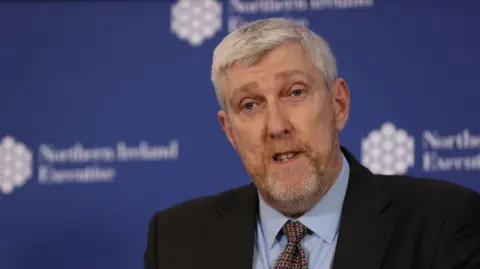 PA Media John O'Dowd has grey hair parted to the side and a thin grey/red beard and is wearing a suit 