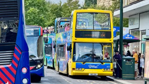 LDRS A yellow open top bus on the road with people queueing to get on and other buses around.