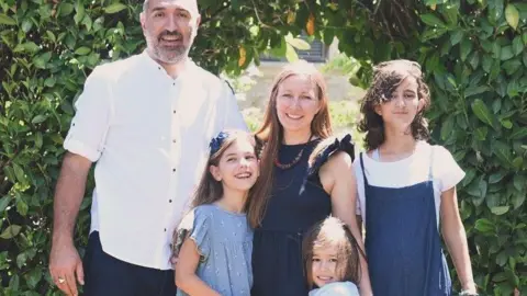 Submitted A family of five smiles for a photo taken outside, with a lush green bushy archway, in a garden. A man, a woman and their three children. One child looks teenage-aged, the others are younger.
