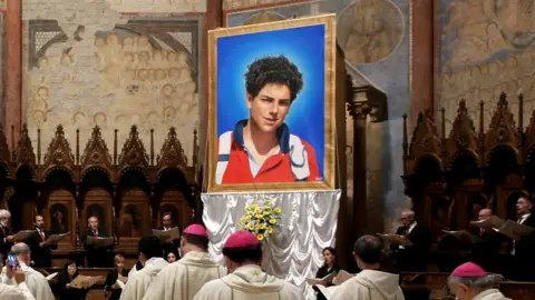 Getty Images A portrait of Carlo Acutis on a platform in a cathedral. He is a teenage boy in a red jacket. There are people wearing Catholic religious attire and the cathedral has gothic architecture.