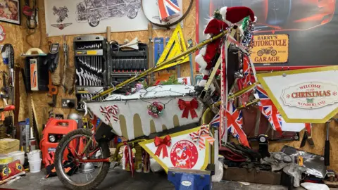 A wheelbarrow covered in Christmas decorations sits on a work bench in a garage. The wheelbarrow's decorations include white felt which looks like dripping snow, red and white bows, baubles, Union Flag bunting, a Santa, and fairy lights.