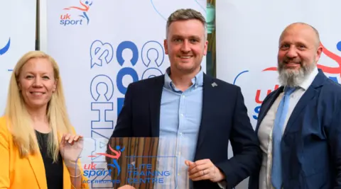 A woman with long blonde hair on the left is wearing a yellow jacket and standing next to a man in the middle with a shirt and dark blue jacket. A man with a grey beard on the right is wearing a shirt, tie and blue jacket.