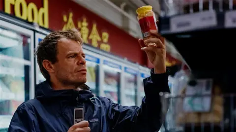 Dan Kelly holding up a jar of pickles and holding a dictaphone in his other hand. Some supermarket fridges are seen in the background.