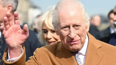 King Charles has grey hair and is smiling. He can be seen waving and is wearing a burgundy coloured coat jacket and light blue tie. A number of figures are in the background