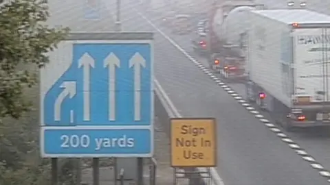 A motorway traffic camera showiing a single line of heavy traffic.