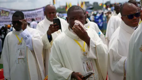 Reuters Four clergymen, in front of a crowd. The one in the centre is wiping sweat from his face with a hankerchief. 