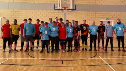 A group of goalball players and volunteers are stood together in front of a set of goals in a sports hall.