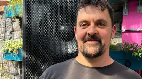 Harry Bradford is standing in a walled garden in front of a black speaker. He is wearing a grey shirt and is smiling.