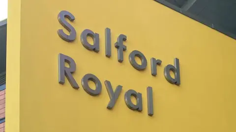 The Salford Royal Hospital sign. It is grey text emblazoned upon a yellow background.