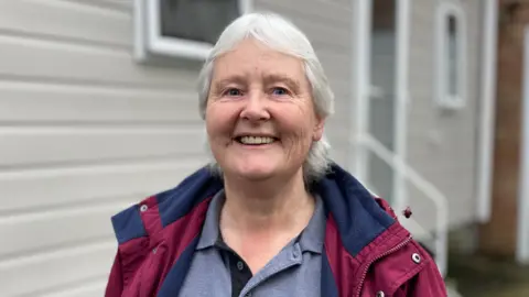 A woman with grey hair and a maroon-coloured coat is standing in front of a static caravan