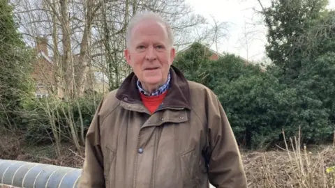 David Chambers stands on the roadside on Market Street in front of the remaining wall by the Kyre Brook. He is wearing a brown jacket, red jumper and chequered shirt.