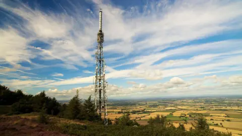 A 200ft tall lattice transmitter mast with communication dishes all up the sides, and a white structure at the top. The mast is set near the summit of a hill, dwarfing surrounding coniferous trees, and towering over a view of patchwork fields for miles into the distance.