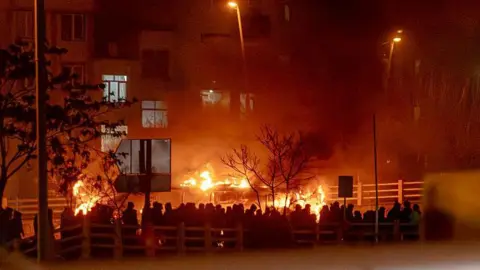 AFP via Getty Images People gather while blocking a street during a protest in Tehran, silhouetted against a burning building.