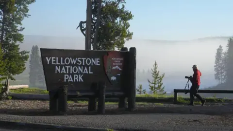 A man holding a camera walks toward a large sign that reads "Yellowstone National Park"