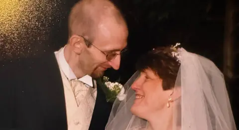 Family photo Jenny and Colin smile and look at each other on their wedding day, Jenny wearing a white veil and Colin in a white waistcoat and shirt and black suit jacket. 