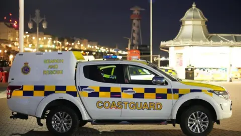 A coastguard vehicle with blue and yellow markings is parked on Brighton seafront at night, with illuminated seaside buildings, streetlights, and a pavilion-style structure visible in the background.