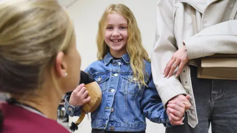 Raoul Hollants A young girl with long blonde hair and wearing a denim jacket is smiling and holding a woman's hand, and a soft toy. In front of them in the foreground a woman in a nurse's uniform kneels and looks up at the child.