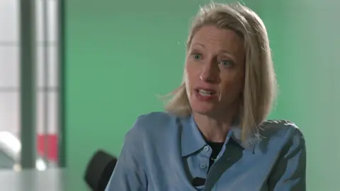Claire Waxman, Victims' Commissioner, she has blonde shoulder length hair, and is wearing a blue shirt, sitting by a table and behind her is a green wall.