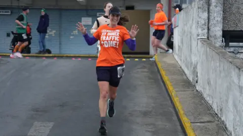 Richard Knights/BBC Runners in motion in a multi-storey car park, with a woman in the foreground running down a ramp. She is wearing an orange tshirt, which has 'Channeling my inner traffic cone' printed on it, in white, and black shorts and baseball cap. Two other female runners are behind her.
