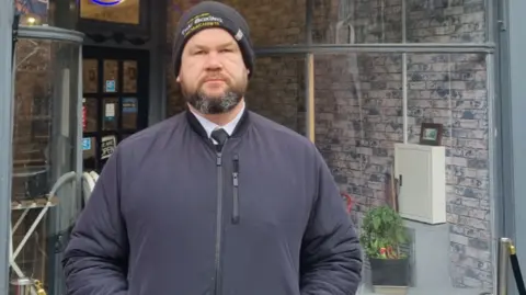 Martin Griffiths, wearing a wool hat and a jacket, standing with his hands in his pockets in front of the High Street Feeder cafe, near where the incident took place.