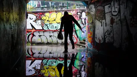 PA Media A person shelters from the rain in the flooded Lawrence Hill underpass in Bristol. They are silhouetted against the wall behind, and are carrying a bag in one hand. Behind them is a wall covered in graffiti, and the person and the graffiti is reflected in the water in the underpass