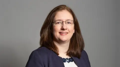 House of Commons Head and shoulders shot of Dr Caroline Johnson. She has dark shoulder-length hair and is wearing glasses and a blue jacket.