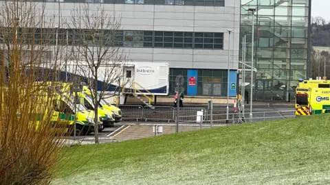 The exterior of Great Western Hospital, taken from a distance. Five ambulances can be seen parked outside, and the picture has been taken from the opposite side of a stretch of grass which has frost on it. There is a large freestanding unit outside the the building, and the main hospital is glad in grey panels with large windows. 