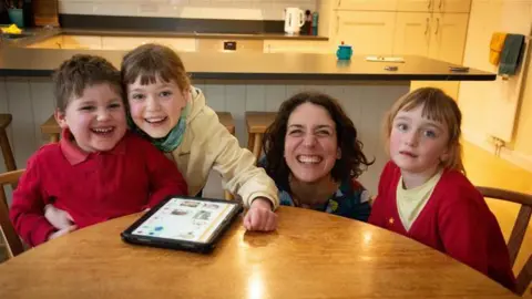 A five-year-old boy, two school-aged girls and their mum are sat around a dining table in their kitchen, smiling broadly at the camera. A tablet rests on the table. The boy has short brown hair and is wearing a red polo shirt and red sweatshirt. One of the girls, who has brown tied back hair and is wearing a yellow hoodie, has her arm around him. Next to her is their mum who is crouched down behind the table and has short, dark curly hair. On her other side is the other girl who is wearing a yellow t shirt and red cardigan and has brown tied-back hair.