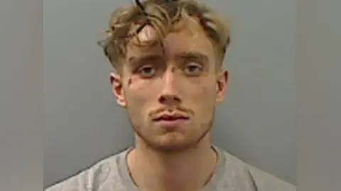 Cleveland Police Mugshot of Callum Howe. He has blonde curly short hair and cuts to his face. He has a tiny moustache and is wearing a grey t-shirt.