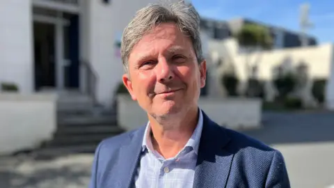 BBC Andy Sloan has grey and black hair. He is wearing a blue suit jacket and a blue and white checked shirt. 