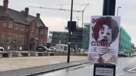 A sign that says down with the clown, featuring a picture of Ronald McDonald, is attached to a traffic light pole. A street is visible in the background