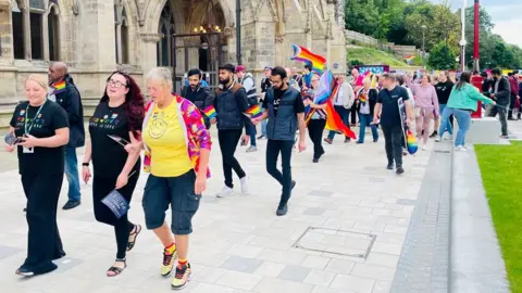 Rochdale Council A line of people dressed in colourful clothing and carrying rainbow flags walk in a parade outside Rochdale's stone-built gothic town hall