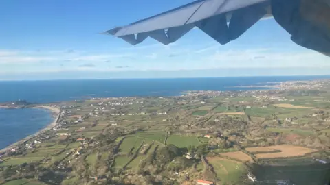 The picture shows the left wing of a white plane from the passenger window. The plane is flying over the west coast of Guernsey.