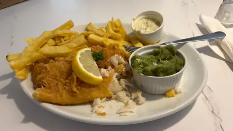 A plate of battered fish and chips, with a slice of lemon, a pot of mushy peas and tartare sauce. A fork is resting on the side of the plate. 