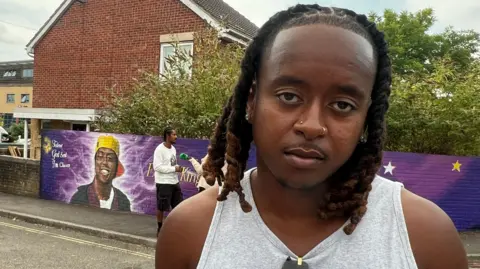 Elliot looks at the camera with a serious expression, the mural visible behind him. He wears a grey vest top, and has dark hair in shoulder-length twists as well as two gold nose piercings and pierced ears. 