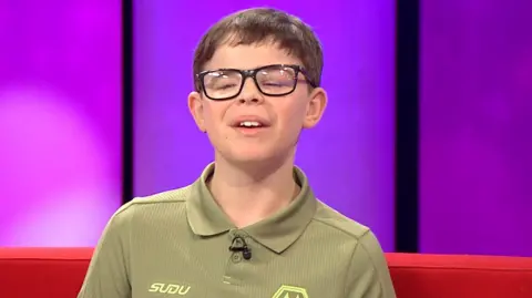 A young boy with short brown hair and wearing glasses and a green shirt is sitting on a red sofa in a TV studio. There is a purple wall behind him.