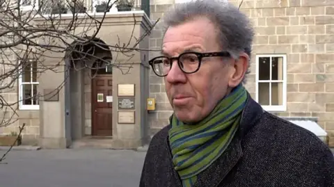 William Tate stands in front of a building with a stone facade. He is wearing glasses, has medium-length dark grey hair and is wearing a dark coat and a green and blue striped scarf.