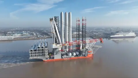 A massive offshore wind installation ship carrying turbine components sails along a river near an industrial port.