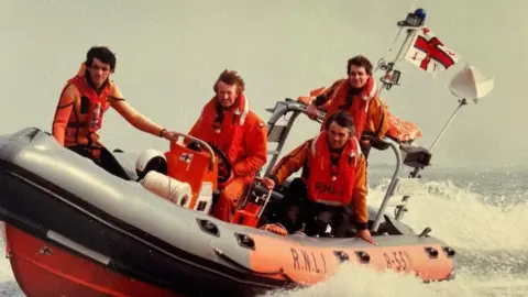 Richard Spindler An old photograph of Richard at the helm of an RNLI speedboat with three other rescuers. They are speeding through a body of water.