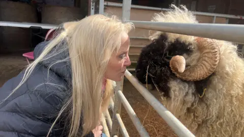 BBC/Emma Kaye A woman in a grey jacket who has long blonde hair, looks through the bars at a sheep which is looking back at her. 