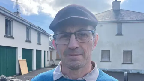 BBC Sean Faloon standing in front of Clonduff parochial house on a sunny day in spring 2025. He has short, greying dark hair and is wearing a dark flat cap, glasses and a blue jumper over a polo shirt.