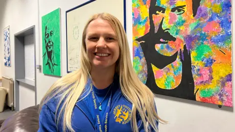 Jo has long blonde hair. She is wearing a blue hoodie with Jersey Youth Service yellow branding on the front.