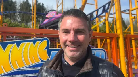 A man standing in front of a colourful amusement‑park ride, with bright yellow and blue coaster tracks and a pink‑and‑purple car behind him, smiling while wearing a dark jacket on a sunny day with trees in the background.