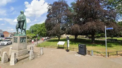A town centre park with large trees with a pavement around the outside, and a small black railing separating the pavement and the park. There is an information board alongside the railing. A statue of the author John Bunyan coated in green patina can be seen to the left of the picture on a stone plinth, with small concrete posts at the base. There is a road to the left of the statue.