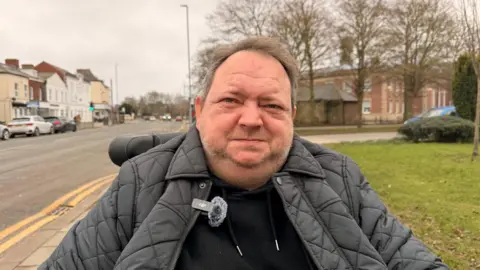 A man sitting in a wheelchair on a path looking at the camera. He is wearing a black hoodie and a black padded jacket. He has short brown hair and stubble. Large buildings can be seen on either side of a road, with cars parked by the side of the road.